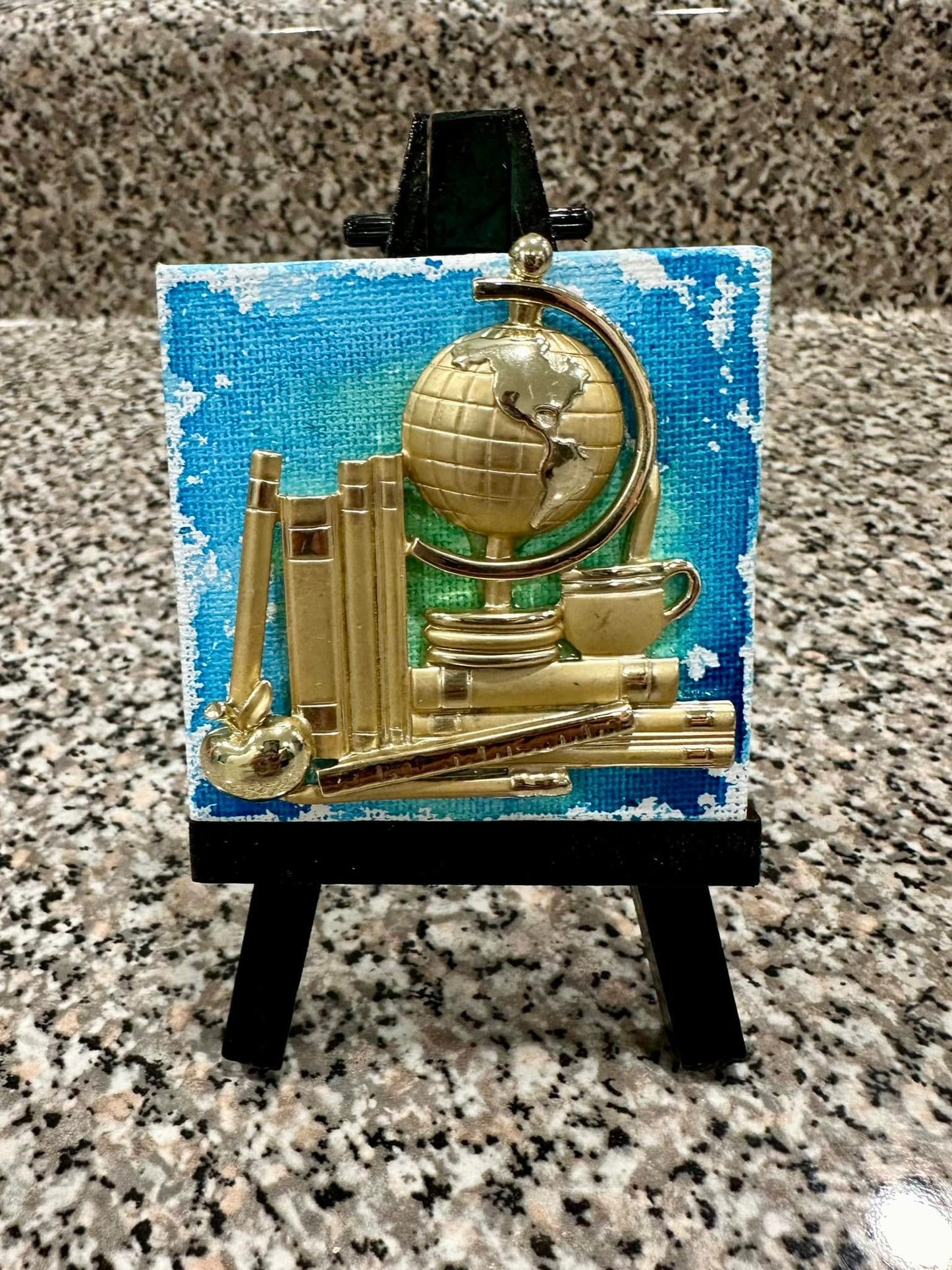 Decorative brooch with a globe and books on a blue and green background, placed on a black mini-easel