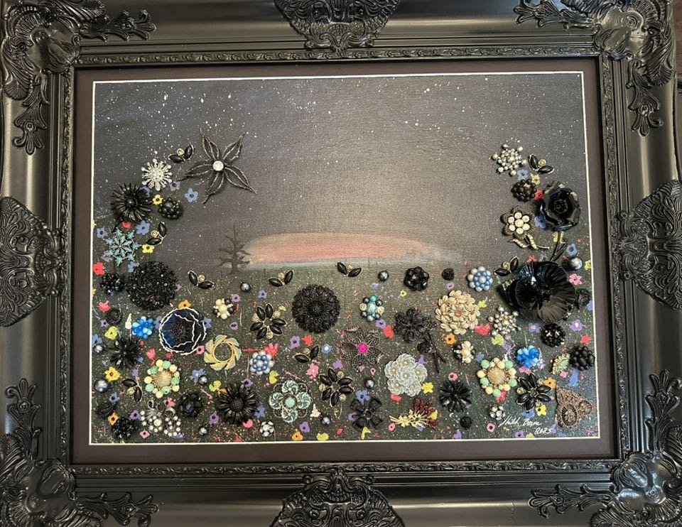 Decorative artwork with vintage jewelry, flowers and a landscape in an ornate black frame.