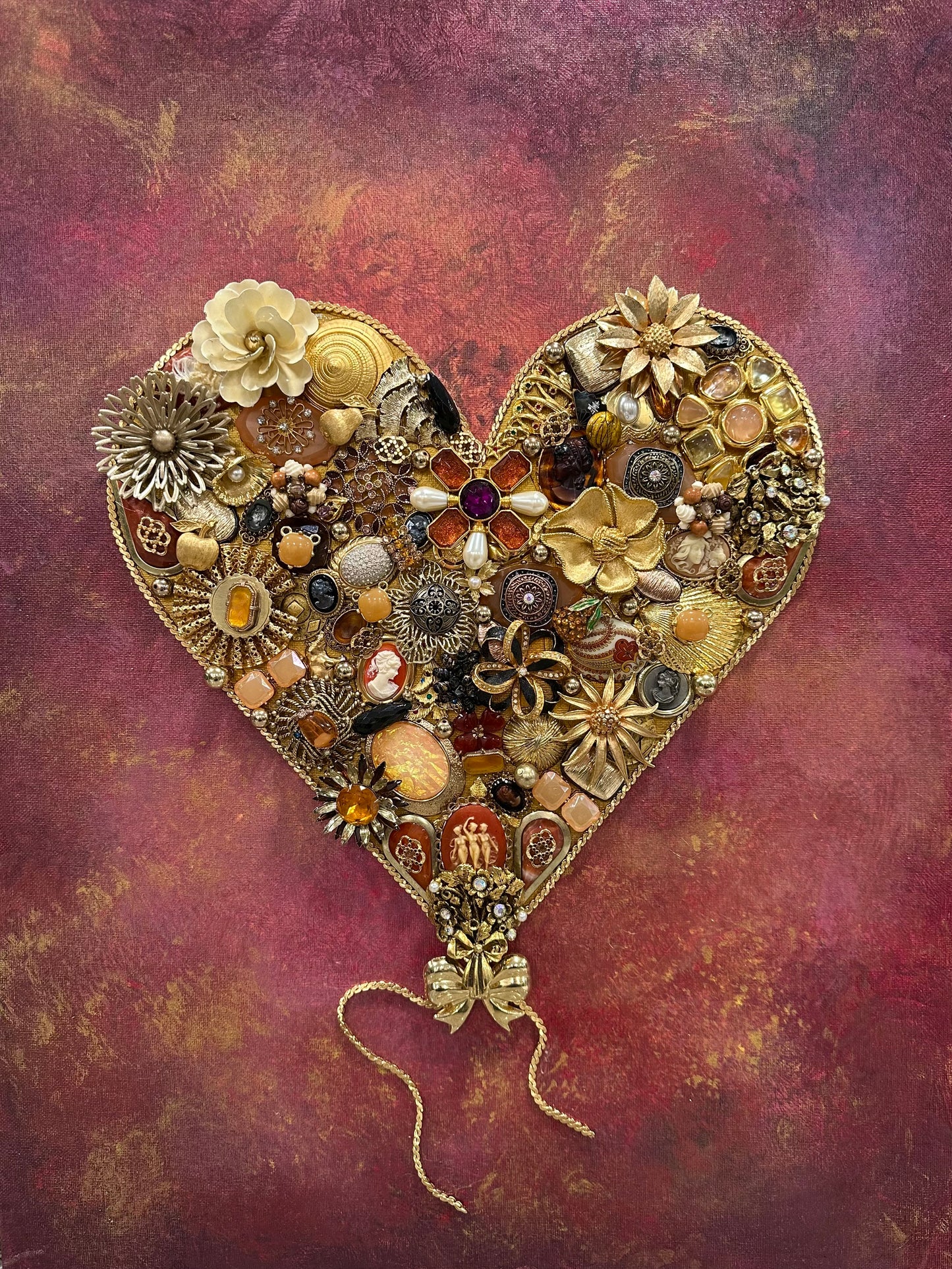 Heart-shaped decorative item made of golden vintage jewelry on a textured background with pink, purple, red, yellow, gold, and black features.
