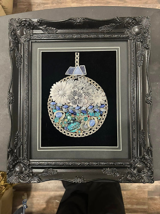 Decorative art piece made of vintage jewelry with floral and gemstone design in a black frame.