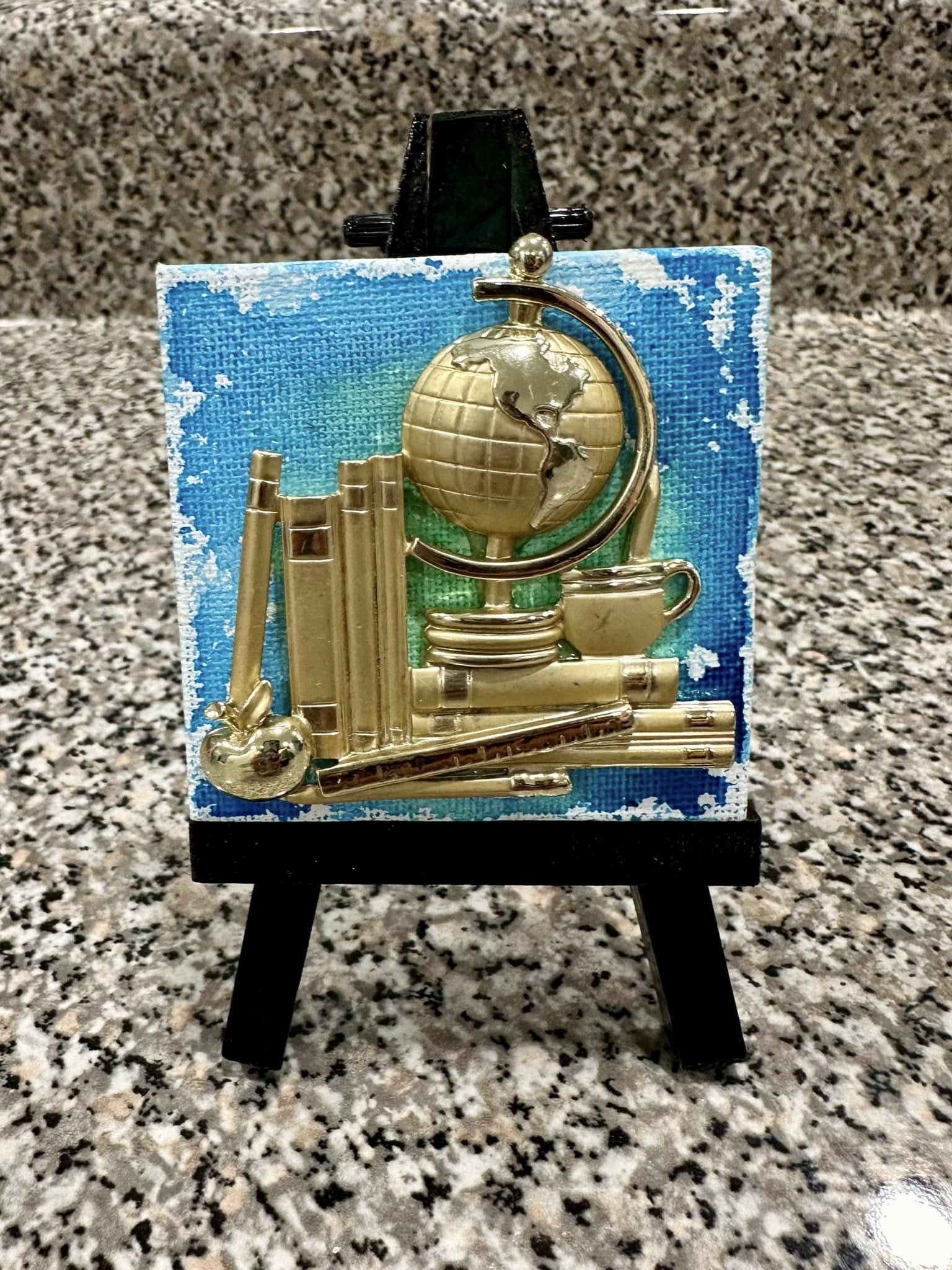 Decorative brooch with a globe and books on a blue and green background, placed on a black mini-easel
