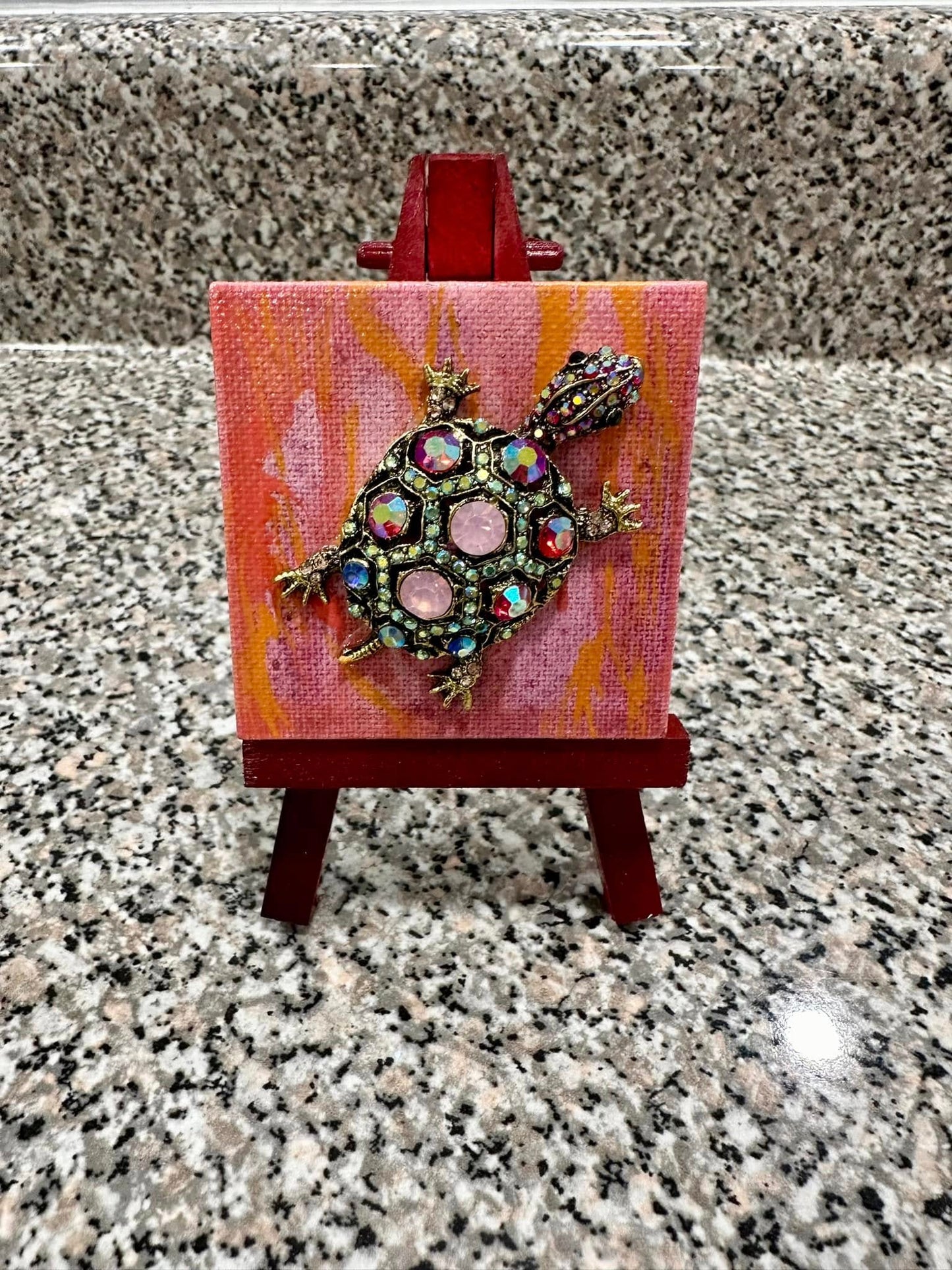 turtle brooch with pink iridescent stones on a red, pink, and orange background on small easel 