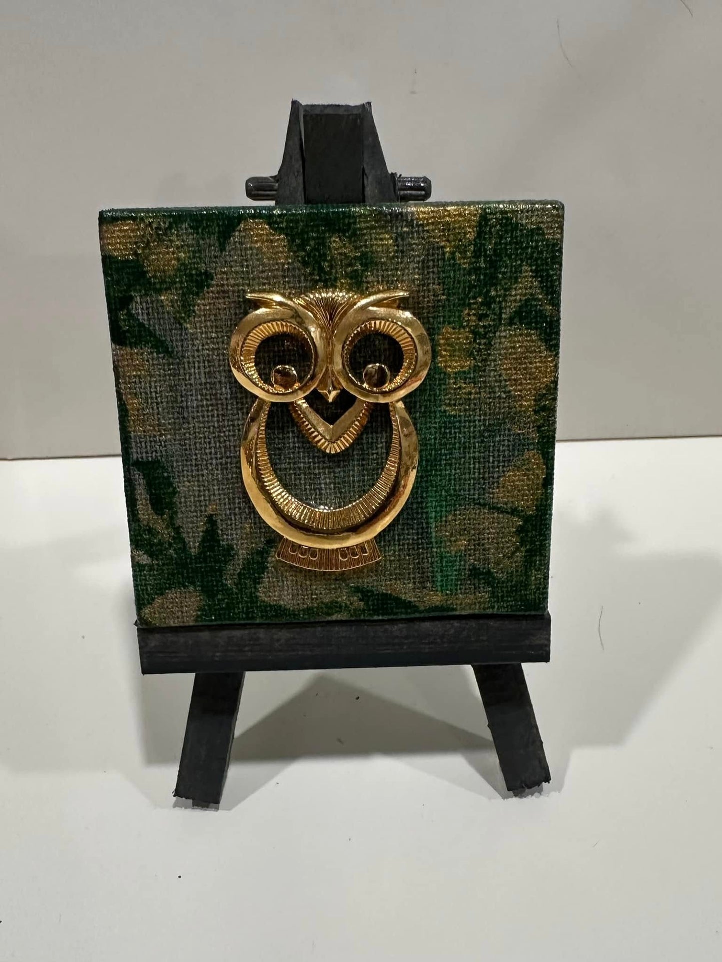 Decorative golden owl  jewelry piece with green and gold background on a mini easel.