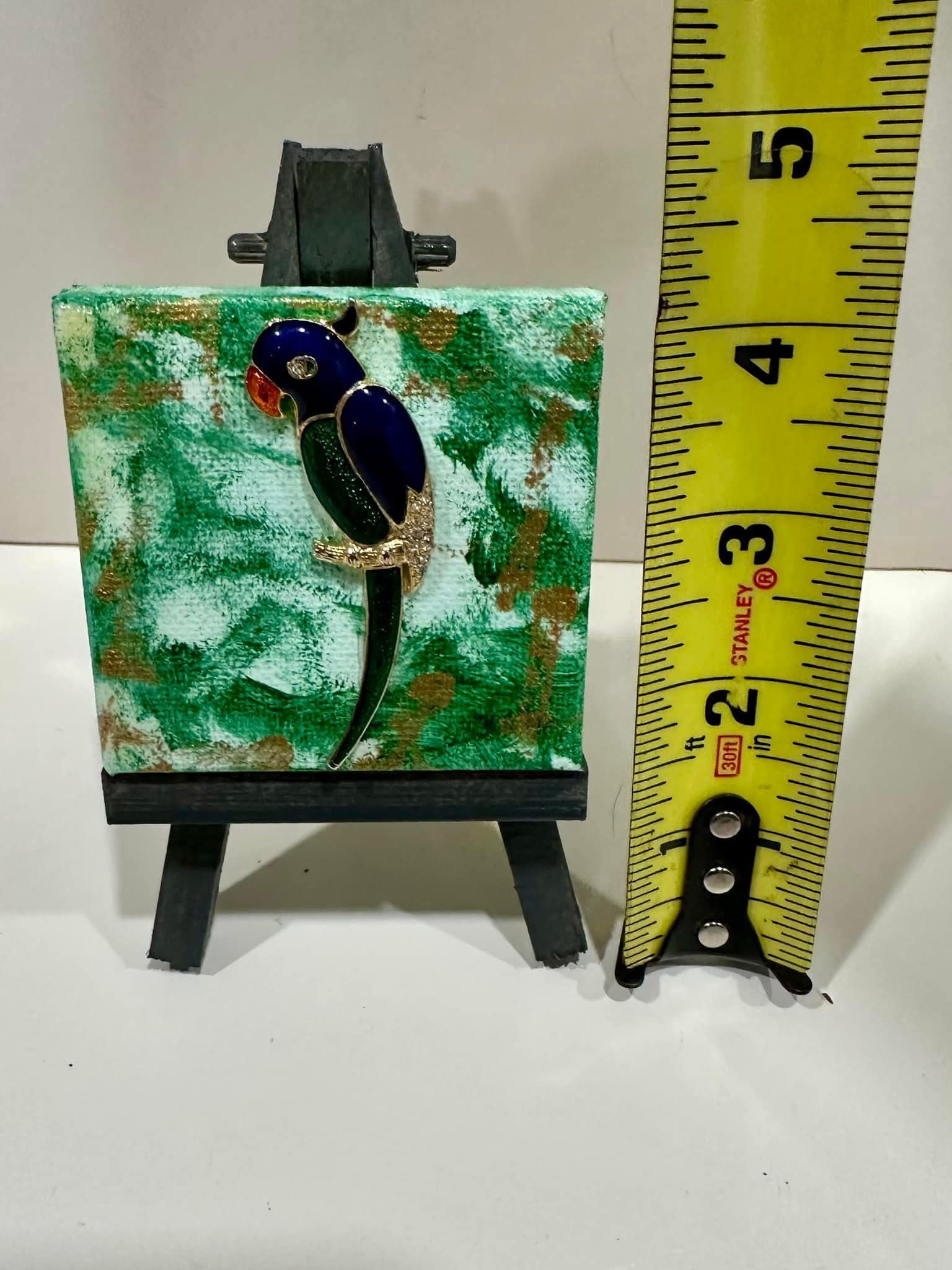 Small brooch of a parrot with painted background on mini easel with a ruler for scale.
