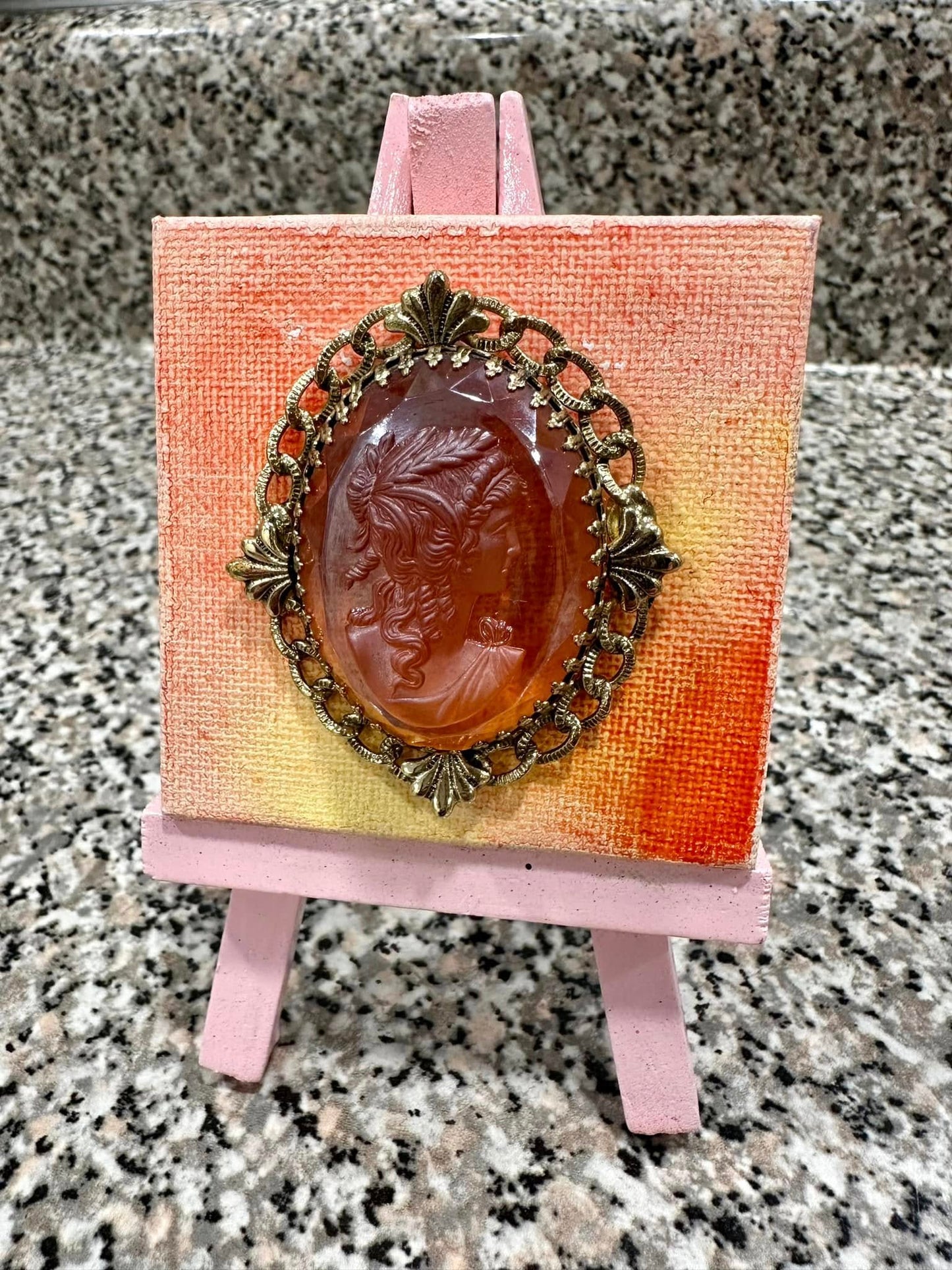 Vintage-style orange cameo brooch on a small easel with a yellow and orange original art background