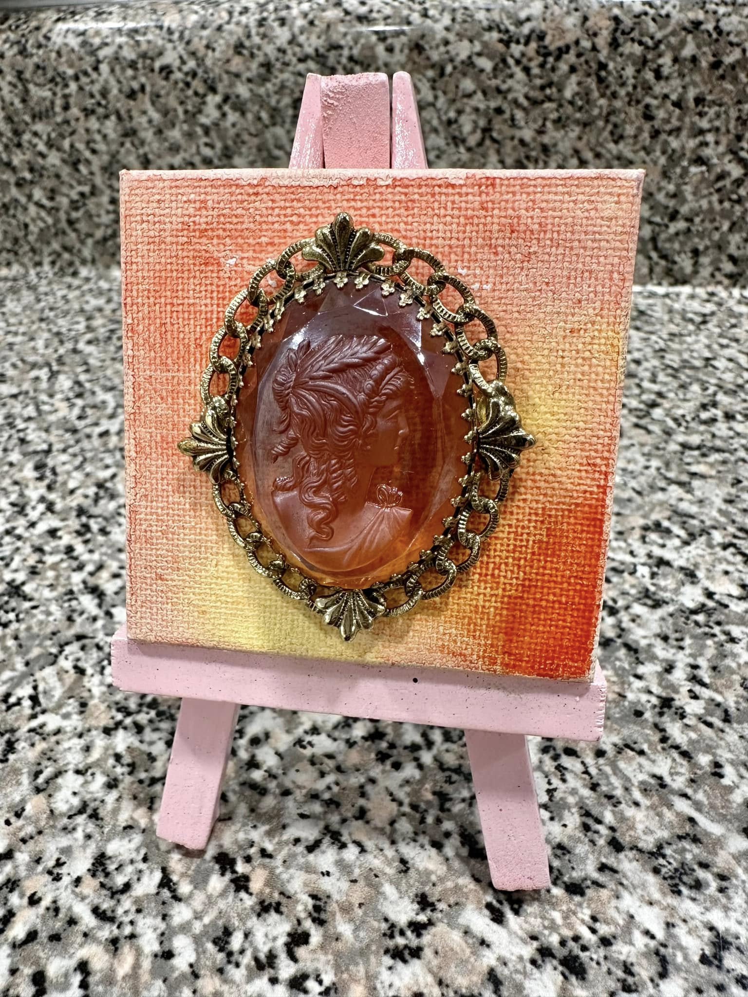 Vintage-style orange cameo brooch on a small easel with a yellow and orange original art background