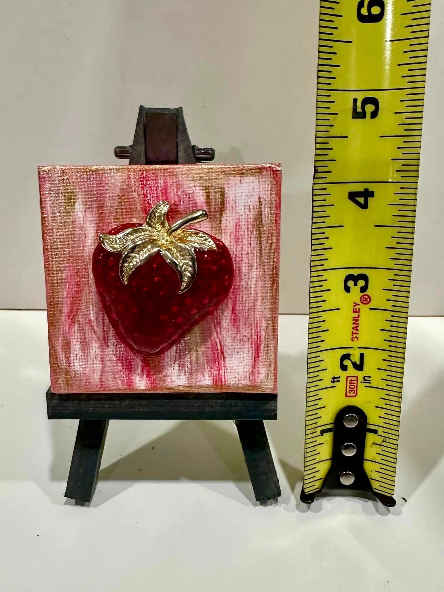 Small red strawberry brooch with gold leaf on a red and gold background, displayed on a mini easel 